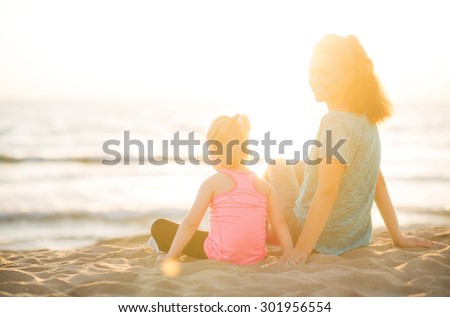 Happy young mother and daughter, seen from behind, are taking a little time to sit and play in the sand at sunset on the beach after a good run together.
