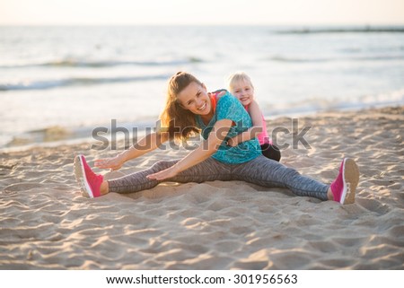 Come on, Mama, you can stretch, you can do it. Here, a little girl helps her mother get more reach as she stretches her arms towards her feet. They are sitting on the sand near the water at the beach.
