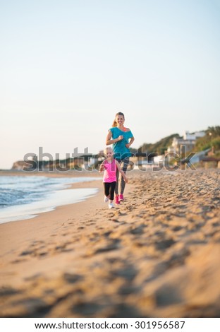 A happy, fit young mother is running behind her daughter on the beach at sunset, along the water. Her daughter is running fast and is delighted to be running faster than her mother.