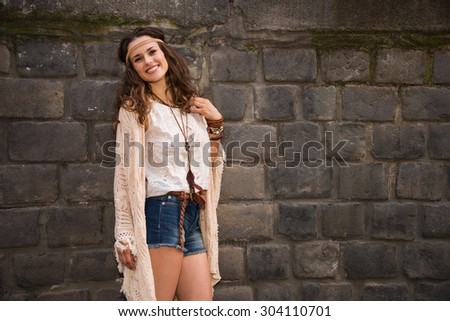 Longhaired hippy-looking young lady in jeans shorts, knitted shawl and white blouse stands near stone wall in old town and smiling