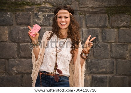 Longhaired hippy-looking young lady in jeans shorts, knitted shawl and white blouse standing near stone wall and showing victory gesture