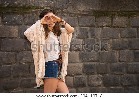 Longhaired hippy-looking young lady in jeans shorts, knitted shawl and white blouse stands near stone wall in old town and hand hearting