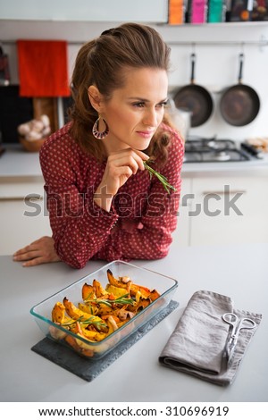 An elegant woman cook leans against the kitchen counter, thinking about the next ingredients. In her hand, she holds a sprig of rosemary. On the counter, hot roasted pumpkin is cooling.