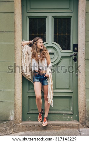 Happy longhaired hippy-looking young woman in jeans shorts, knitted shawl and white blouse standing outdoors against olive wooden door