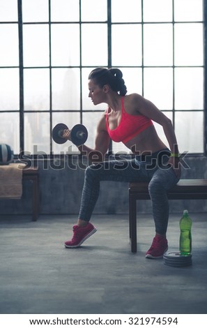 A fit, sporty young woman is sitting in profile on a bench in profile, lifting weights with one hand, while resting her elbow on her knee.