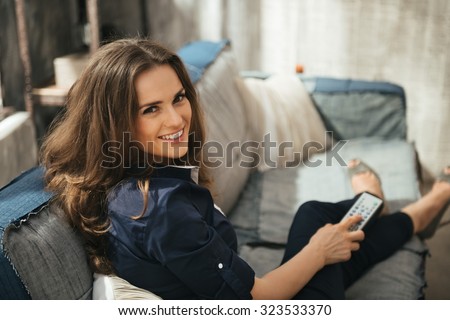 Smiling longhaired woman with TV remote control in hand relaxing on sofa and watching tv in loft apartment. Urban chic loft decoration details and window. Modern lifestyle concept.