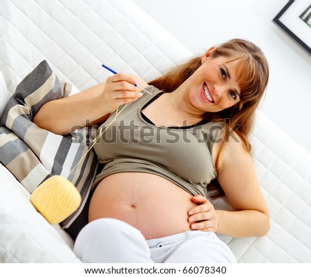 Smiling beautiful pregnant female sitting on sofa and knitting for baby.