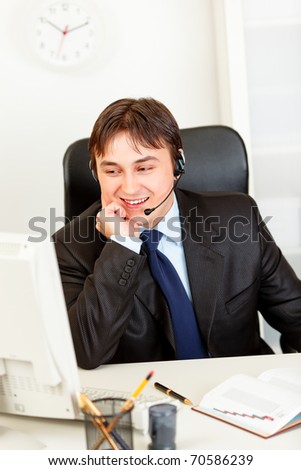 Smiling  business man with  headset sitting at office desk and looking at computer monitor