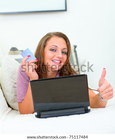 Smiling pretty woman lying on sofa at home with laptop and  credit card and showing  thumbs up gesture