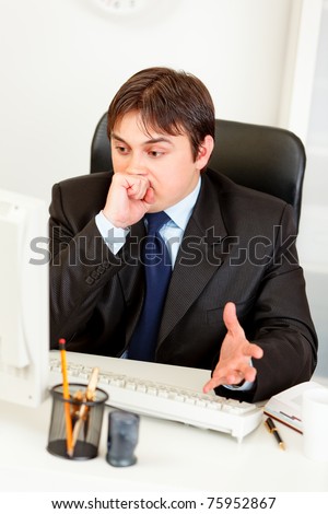 Confused business man sitting at office desk and surprisingly looking at computer monitor