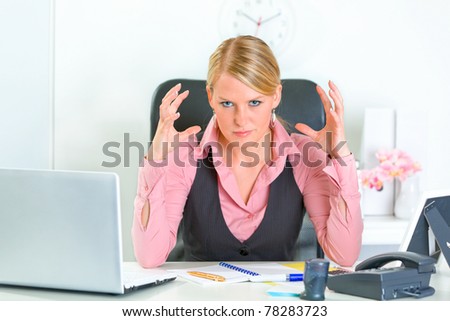 Angry modern business woman sitting at office desk and holding hands near head
