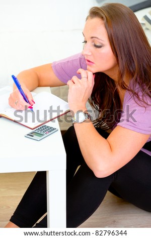 Pensive lovely woman sitting on floor at home and making notes in diary