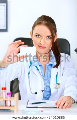 Authoritative medical doctor woman sitting at office table and holding blank business card