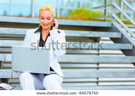 Smiling modern business woman sitting on stairs talking on mobile and looking in laptop