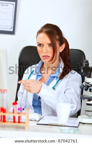 Confused doctor woman sitting at table and pointing finger in monitor