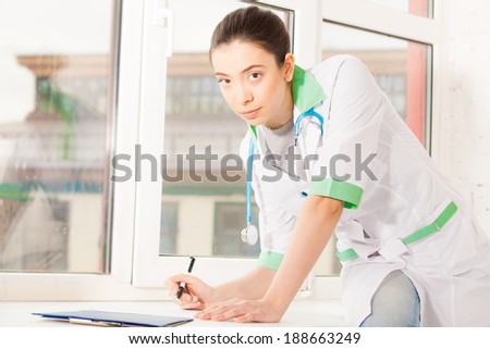 Young doctor sitting on window sill and writing