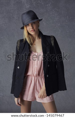 beautiful young blond woman with messy hair in a black blazer, wool hat and rompers on grunge studio background