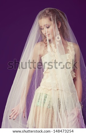 Portrait of a beautiful bride with long curly hair wearing lace dress over shorts standing under tulle veil on purple studio background