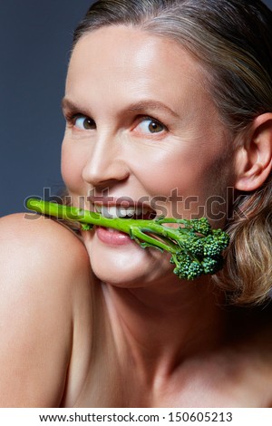 beautiful fourty year old woman with natural makeup and healthy skin texture on blue gray studio background holding a broccoli in her mouth