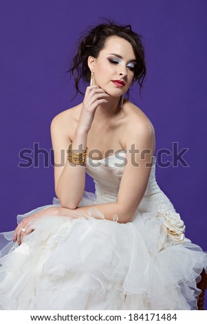 Beautiful young bride in white dress wearing gold accessories sitting in white dress against purple studio background