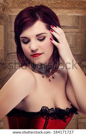 young beautiful woman with red hair wearing red satin and lace corset on architectural background