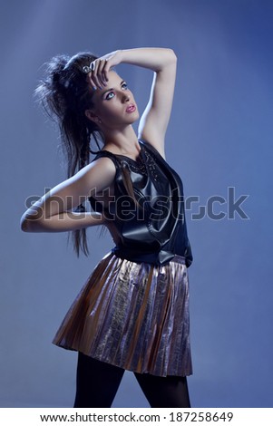 Rocker model girl with long hair in punk hairstyle, wearing black leather top and gold skirt on blue studio background