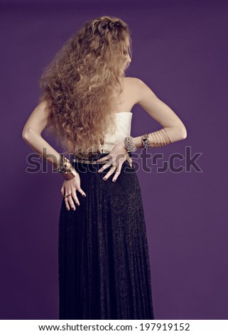 Portrait of a young beautiful woman with long blond curly hair with gold bracelets and rings, standing with her back in a long black skirt against purple studio background