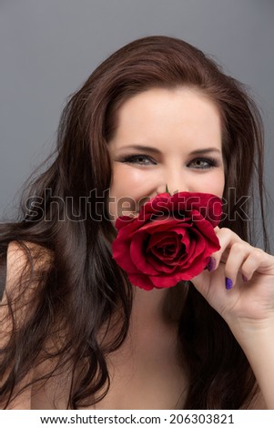 Portrait of a beautiful young brunette woman. Wearing long loose curly hair, posing with a red flower. Against grey studio background. Spa concept