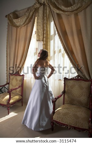 Beautiful bride in silk grey & white dress next to sunlit window with heavy curtains, natural light