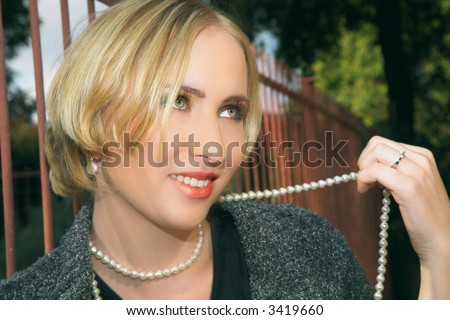 Blond woman with short hair playfully holding string of pearls and smiling, outside