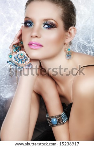 Blond woman with long false lashes holding pearl and blue stones golden jewelry with dreaming expression