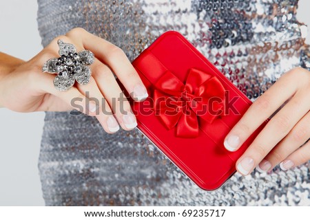 red gift box with satin bow in hands of a young woman wearing a large flower ring and a silver sequin dress .