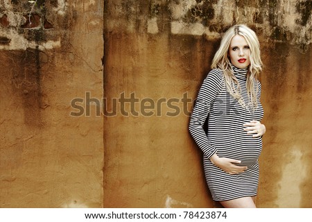 Beautiful eight months pregnant blond woman with curly hair in a black and white stripes fashion mini dress against a grunge wall background .