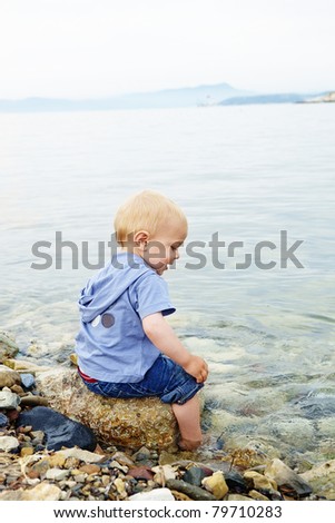 Little blond one year old boy sitting on a rock with his feet in the sea water on a warm summer evening