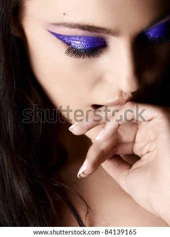 Beautiful young Italian woman with artistic purple eyeshadow and long hair looking down and biting her hand, shot in low key.