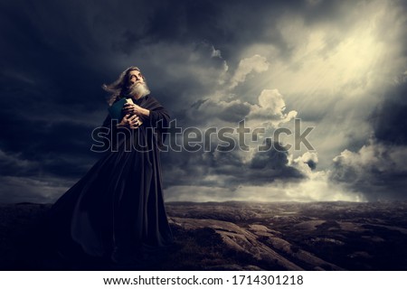 Monk Holding Bible Looking Up to God Sky Light, Old Priest in Black Robe in Storm Mountains