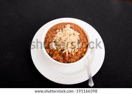 A white bowl of chili con carne with beans served over rice on a black background