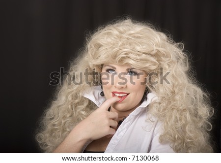 A model wearing a blonde wig and posing for the camera with finger in mouth, looking up
