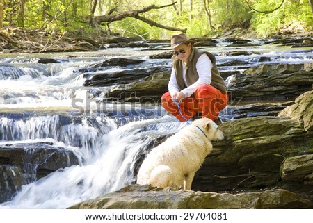 An hispanic man in hat, sunglasses, and orange pants in the river with his dog