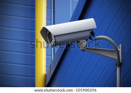surveillance camera on blue wall next to yellow pipe