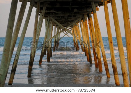 Underneath view of a wooden pier with dusk sunset light shimmering off the poles and water of the ocean\'s edge.