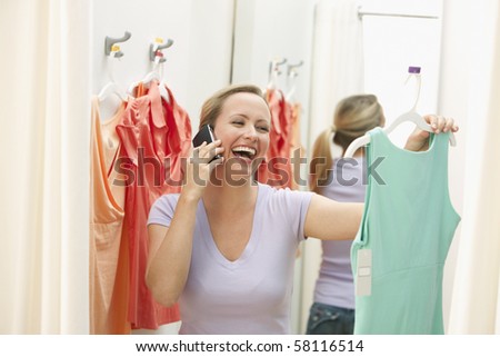A young woman is holding up a dress in a store while talking on a cell phone.  Horizontal shot.