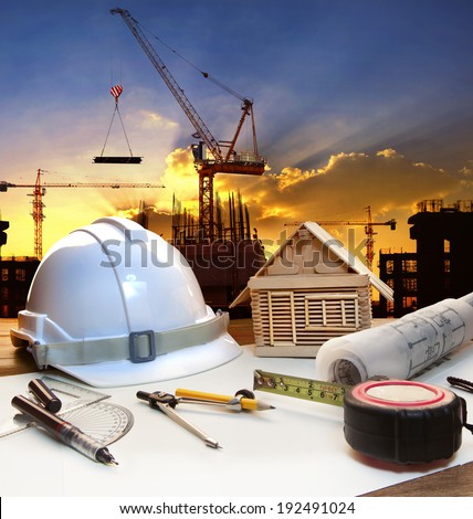 engineer working table plan, home model and writing tool equipment against building construction crane with evening dusky sky