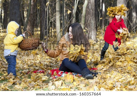 Mother with two children sits on an edge of the autumn wood, collecting leaves. The girl in a red coat jealously looks at the brother