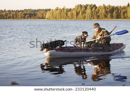 The man in a camouflage floats by the boat on the lake in Karelia with two hunting dogs. One dog looks out for something in water, being going to jump. A back hunter has a shot-gun.