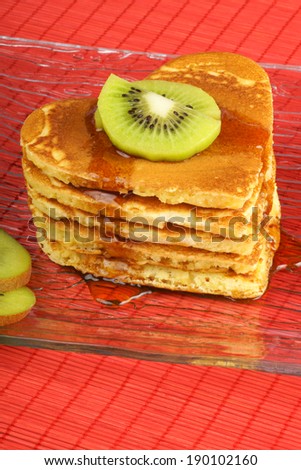 Stack of heart shaped pancakes with syrup and kiwi fruit on a transparent glass dish over a red background. A perfect breakfast for Valentine\'s Day