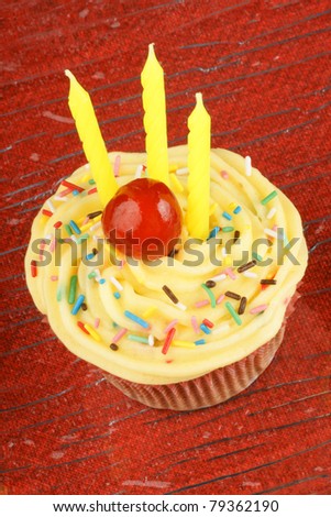 Birthday cupcake with lemon butter-cream, sprinkles, candied cherry and three candles. Over a red background. Selective focus.