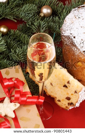 Panettone the italian Christmas fruit cake, a glass of spumante (champagne) and a Christmas present under the Christmas tree, over a red background. Selective focus.