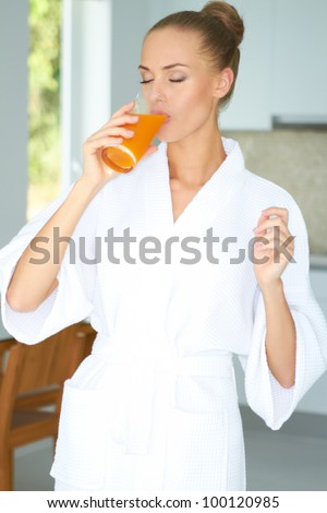 Woman in a white bath robe with her eyes closed enjoying a large glass of fresh orange juice in the morning for breakfast