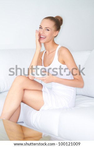 Gorgeous woman with a beautiful smile sitting cross legged on a white sofa drinking coffee